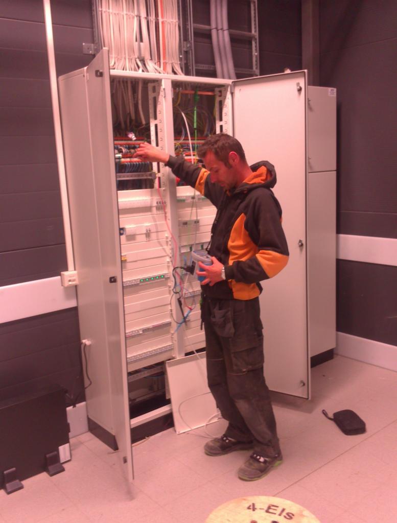 Mężczyzna w roboczym ubraniu sprawdza połączenia w otwartej szafie sterowniczej z licznymi kablami i przewodami, używając specjalistycznego miernika.