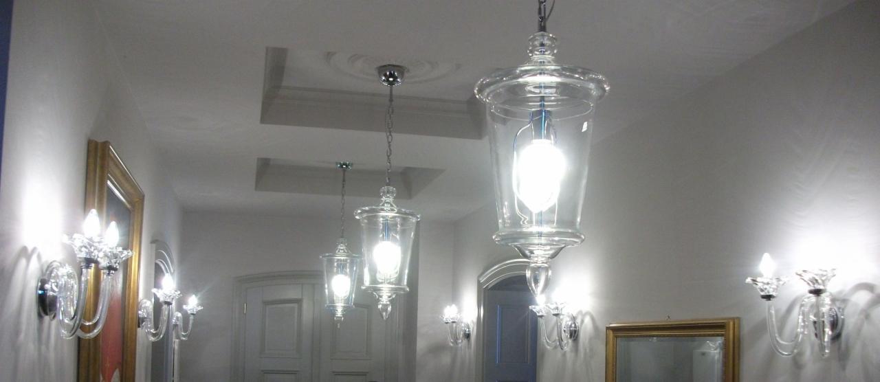 Elegancki korytarz z trzema szklanymi lampami wiszącymi o kształcie lampionów i kryształowymi kinkietami ściennymi, oświetlony ciepłym światłem.