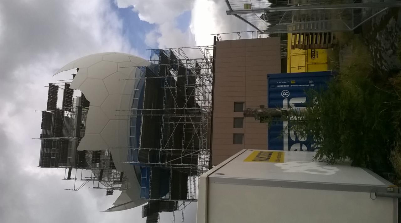 Konstrukcja sferyczna w trakcie montażu, otoczona rusztowaniami, z widocznymi elementami budynku i kontenerami w tle.