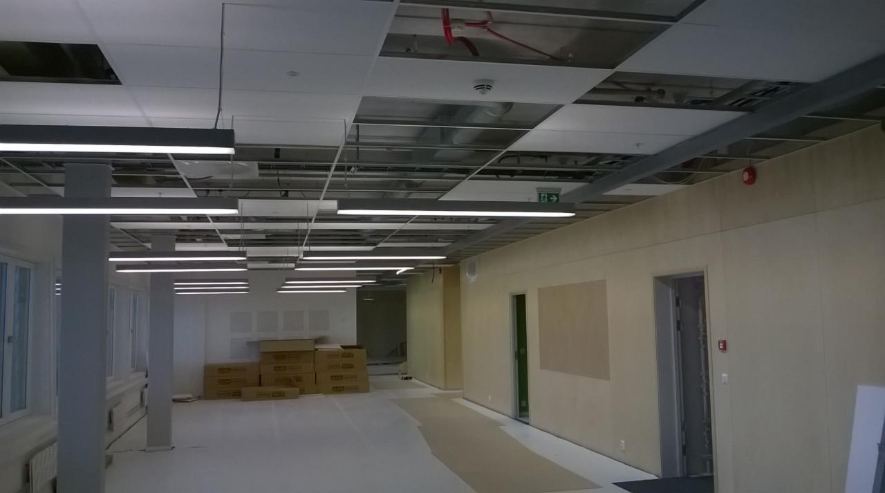 Wnętrze w trakcie wykańczania: białe ściany, podwieszany sufit z widoczną konstrukcją, liniowe lampy LED, kartony na podłodze, częściowo zamontowane panele sufitowe.