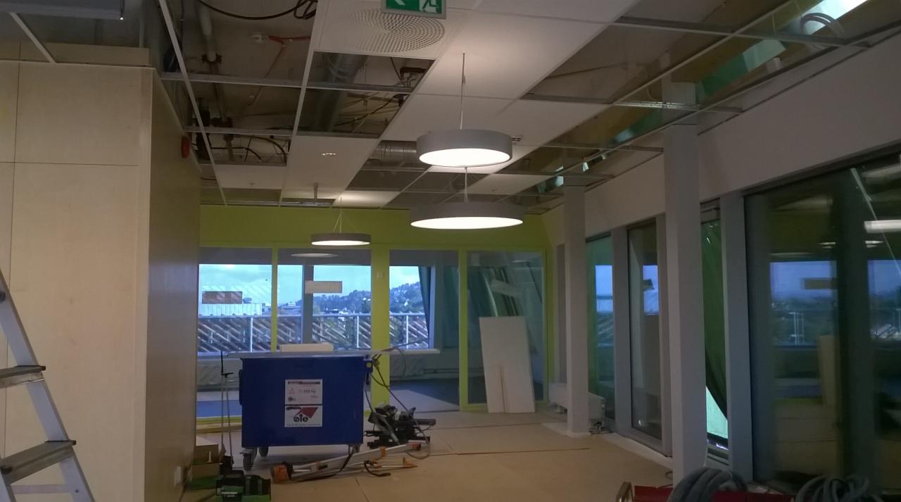 Wnętrze biurowe w trakcie montażu oświetlenia sufitowego, widoczne instalacje podwieszane, narzędzia i materiały budowlane na podłodze, duże okna z widokiem na miasto.