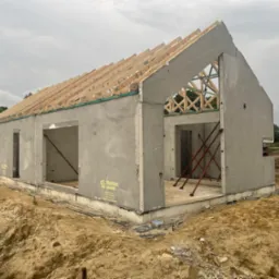 Surowy stan domu z prefabrykatów betonowych z widoczną konstrukcją dachu z drewna i żółtym logo producenta na ścianie, otoczony ziemią.