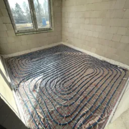 Wnętrze surowego domu z widoczną instalacją ogrzewania podłogowego w postaci niebieskich rurek ułożonych na folii z czerwoną kratką, ściany z białych bloczków, okno z widokiem na drzewa.