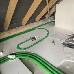 Montaż zielonych rur wentylacyjnych na izolacji poddasza w trakcie budowy, widoczne drewniane elementy konstrukcji dachu i fragment ściany.