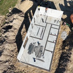 Atrakcyjne Domy Sp. z o.o. - Fundament domu z prefabrykatów betonowych, widok z góry. Na fundamencie widoczne elementy konstrukcyjne i pracownicy w kaskach. Żuraw budowlany w tle.