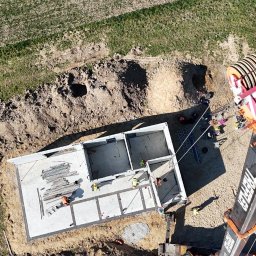 Atrakcyjne Domy Sp. z o.o. - Widok z góry na fundamenty domu w trakcie budowy, z dźwigiem i pracownikami w kaskach, otoczone ziemią i trawą. W tle zaparkowane samochody.