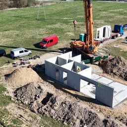 Atrakcyjne Domy Sp. z o.o. - Montaż domu z prefabrykatów: dźwig unosi betonowy moduł, widoczny fundament i kolejne elementy na ciężarówce. Krajobraz budowy, słoneczny dzień.