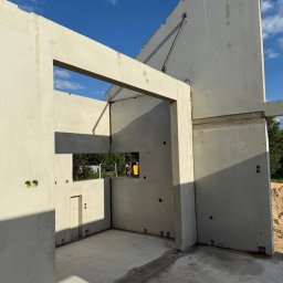 Atrakcyjne Domy Sp. z o.o. - Surowy stan domu z prefabrykatów betonowych w słoneczny dzień. Widoczne otwory okienne i drzwiowe, ślady po szalunkach i elementy złączne. W tle zieleń.