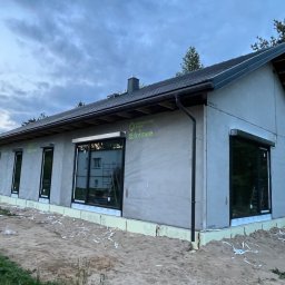 Atrakcyjne Domy Sp. z o.o. - Niski dom parterowy z szarą elewacją w trakcie budowy, z widocznymi oknami z roletami zewnętrznymi i ociepleniem fundamentów. Na elewacji widoczne logo producenta.