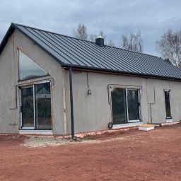 Atrakcyjne Domy Sp. z o.o. - Surowy, parterowy dom z szarą blachodachówką i dużymi oknami, w trakcie wykończenia elewacji. Widoczne ocieplenie wokół okien i czerwona ziemia wokół budynku.