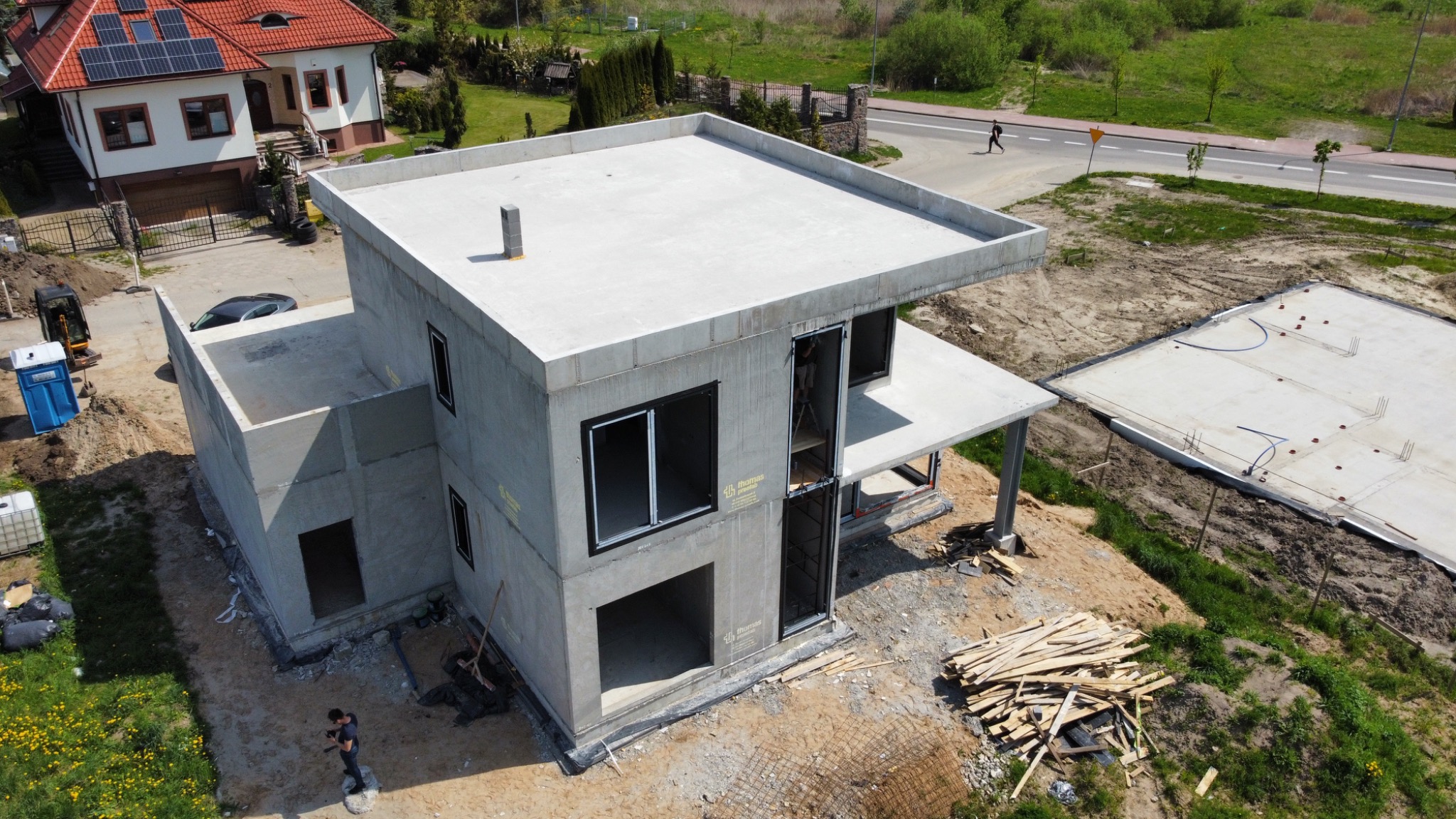 Surowy stan dwukondygnacyjnego domu z betonu komórkowego z płaskim dachem, widok z lotu ptaka, widoczna konstrukcja ścian i otwory okienne, na pierwszym planie stos desek, w tle dom z panelami...