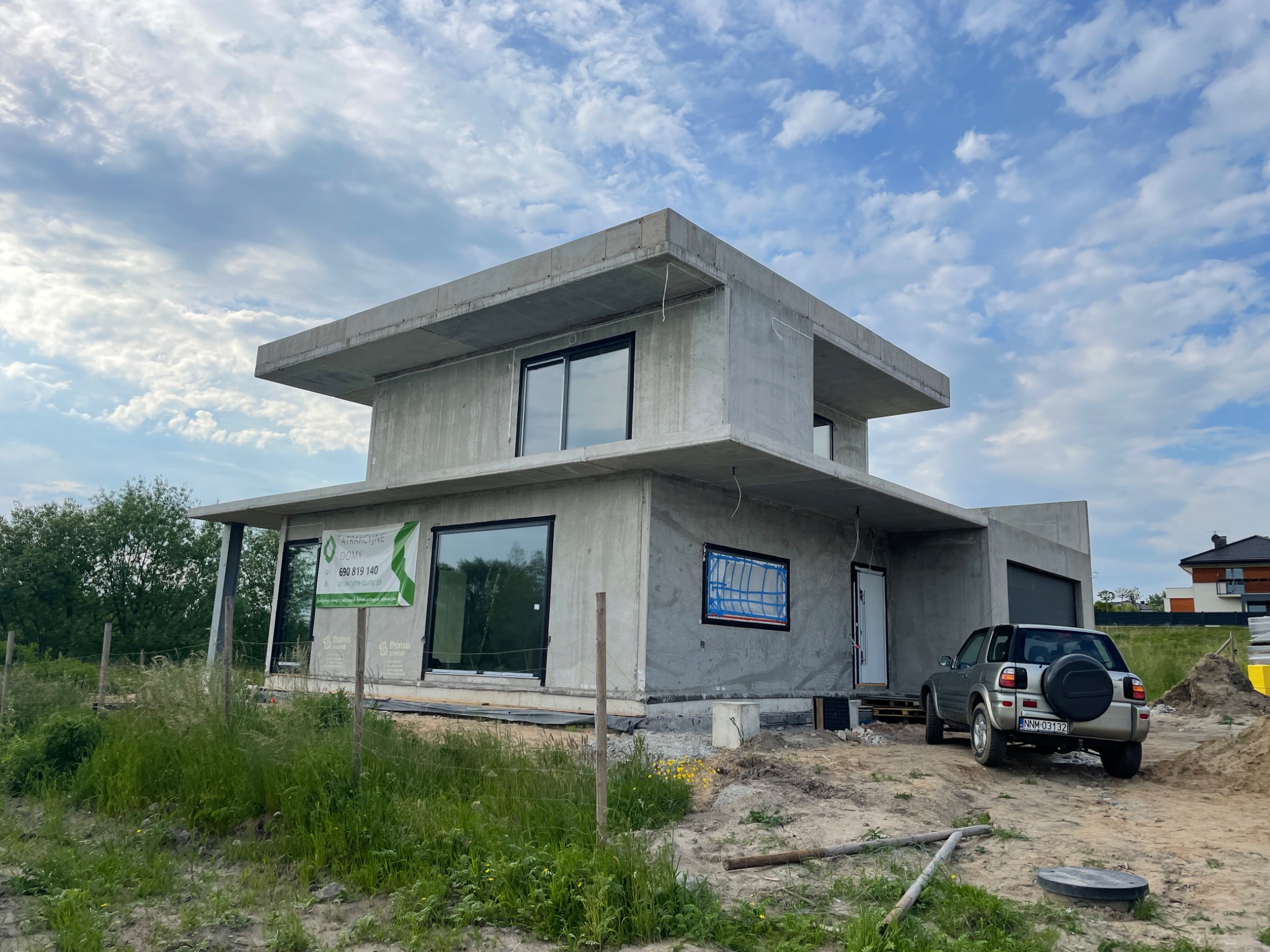 Surowy, dwukondygnacyjny dom w stanie deweloperskim z płaskim dachem, szarym tynkiem zewnętrznym, czarnymi ramami okiennymi i zaparkowanym SUV-em na piaszczystym terenie budowy.