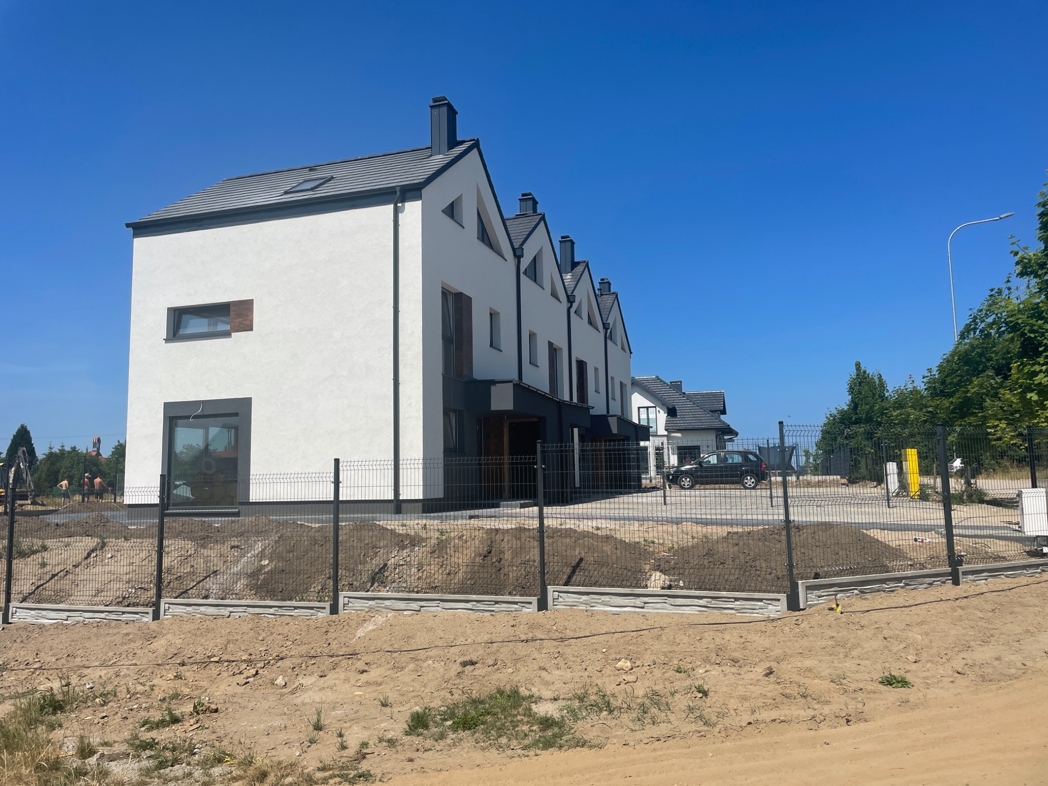 Szereg nowoczesnych, białych domów z ciemnymi dachami i oknami, ogrodzonych metalową siatką na betonowej podmurówce, teren wokół w trakcie prac ziemnych, widoczne maszyny budowlane w tle.