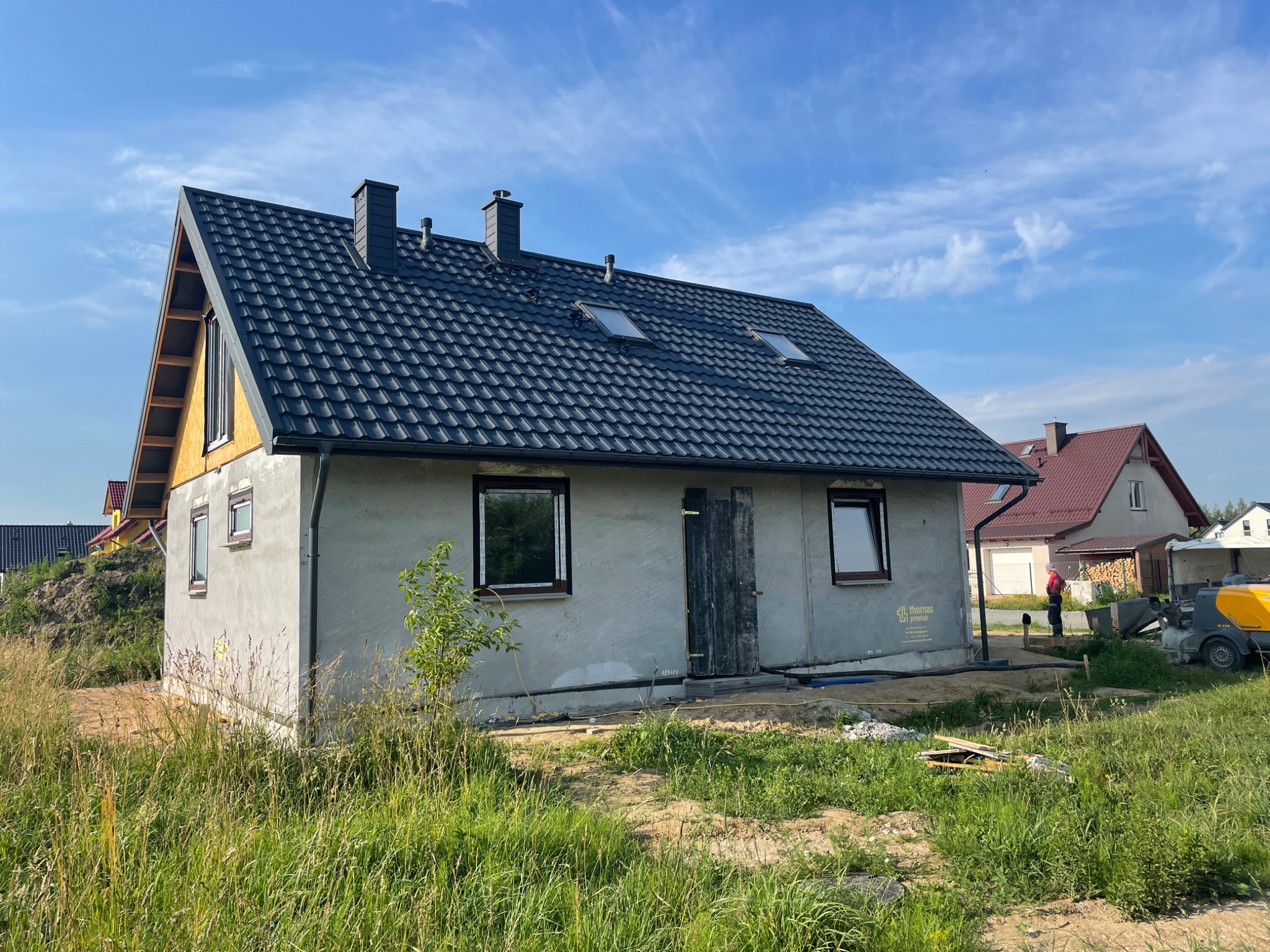 Surowy, otynkowany dom jednorodzinny z ciemną, skośną dachówką i dwoma oknami dachowymi, widoczny z zewnątrz na tle błękitnego nieba, z maszynami budowlanymi w tle.