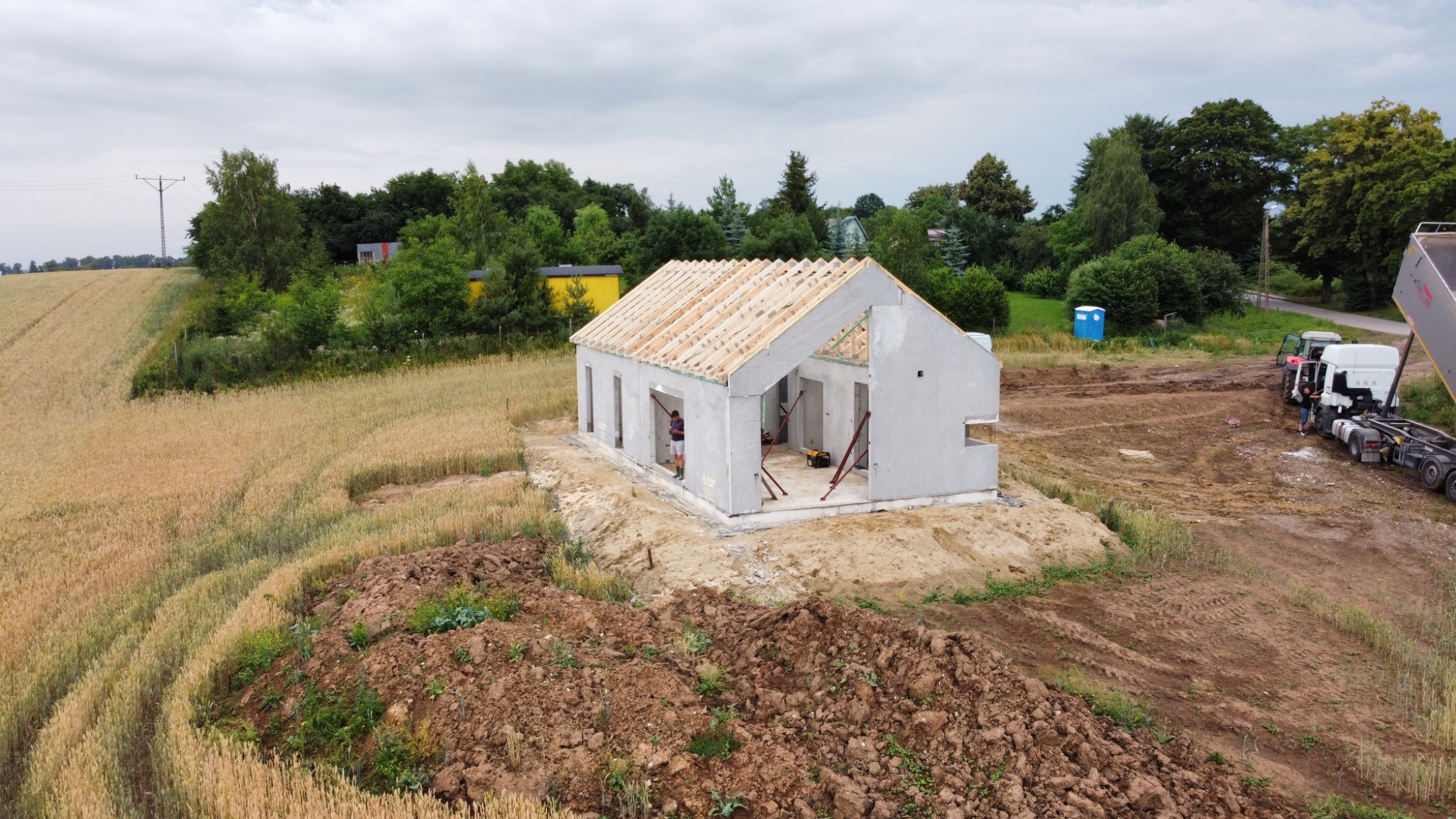 Surowy stan domu z prefabrykatów betonowych z drewnianą konstrukcją dachu w trakcie budowy na działce z polem zboża w tle.