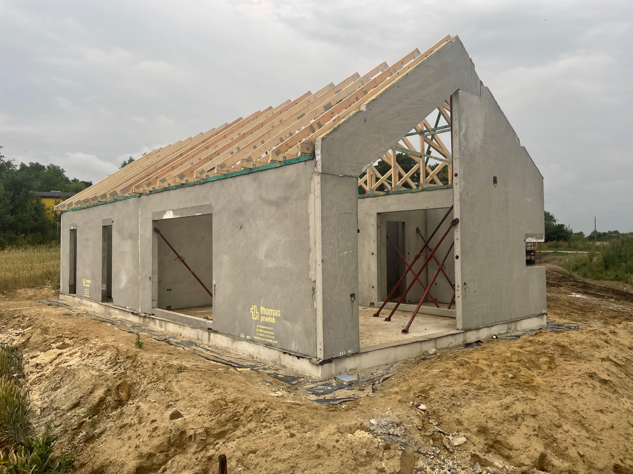 Surowy stan domu z prefabrykatów betonowych z widoczną konstrukcją dachu z drewna i żółtym logo producenta na ścianie, otoczony ziemią.