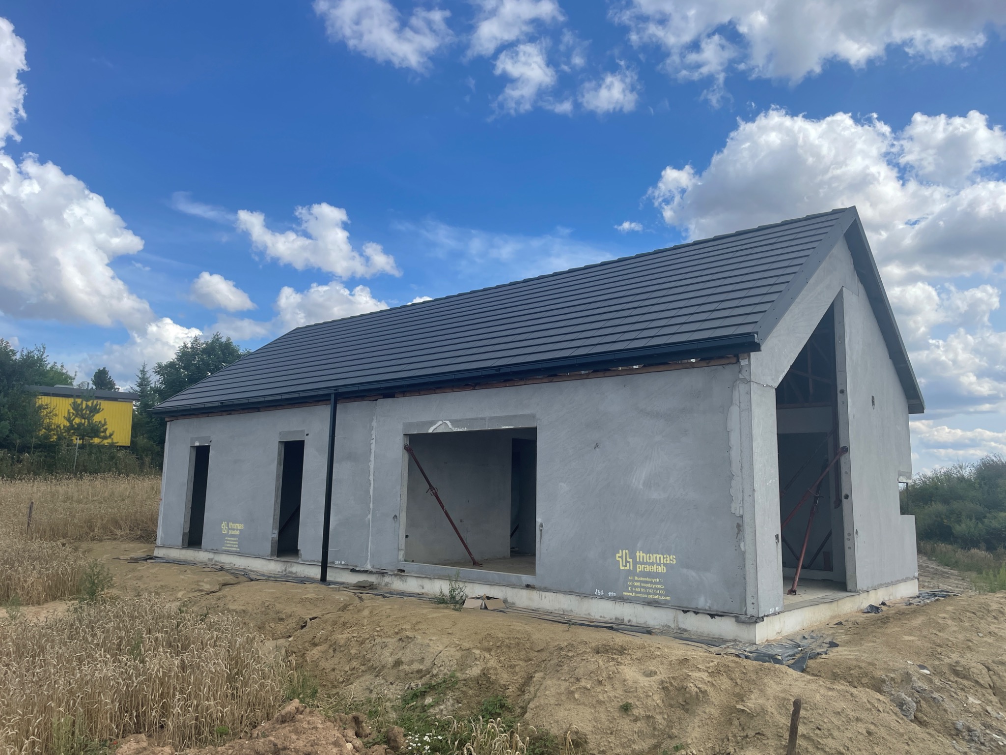 Surowy dom murowany w Gdyni, w trakcie budowy, z widocznym szarym tynkiem zewnętrznym i ciemną dachówką, bez okien i drzwi, otoczony ziemią i wysoką trawą, z logo firmy na ścianie.