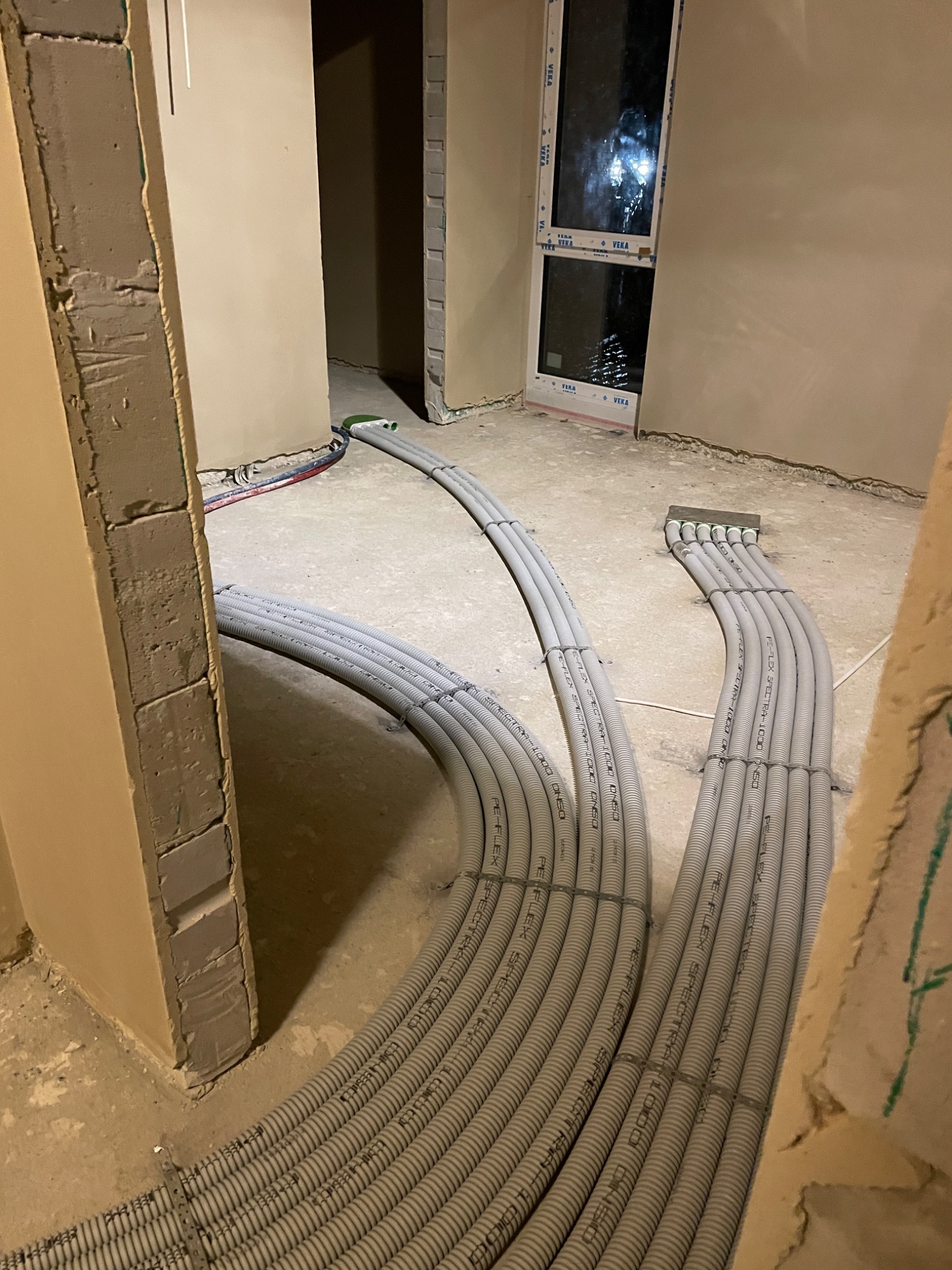 Wnętrze w trakcie prac instalacyjnych: rury instalacyjne ułożone na surowej podłodze, prowadzące do ściany z oknem.