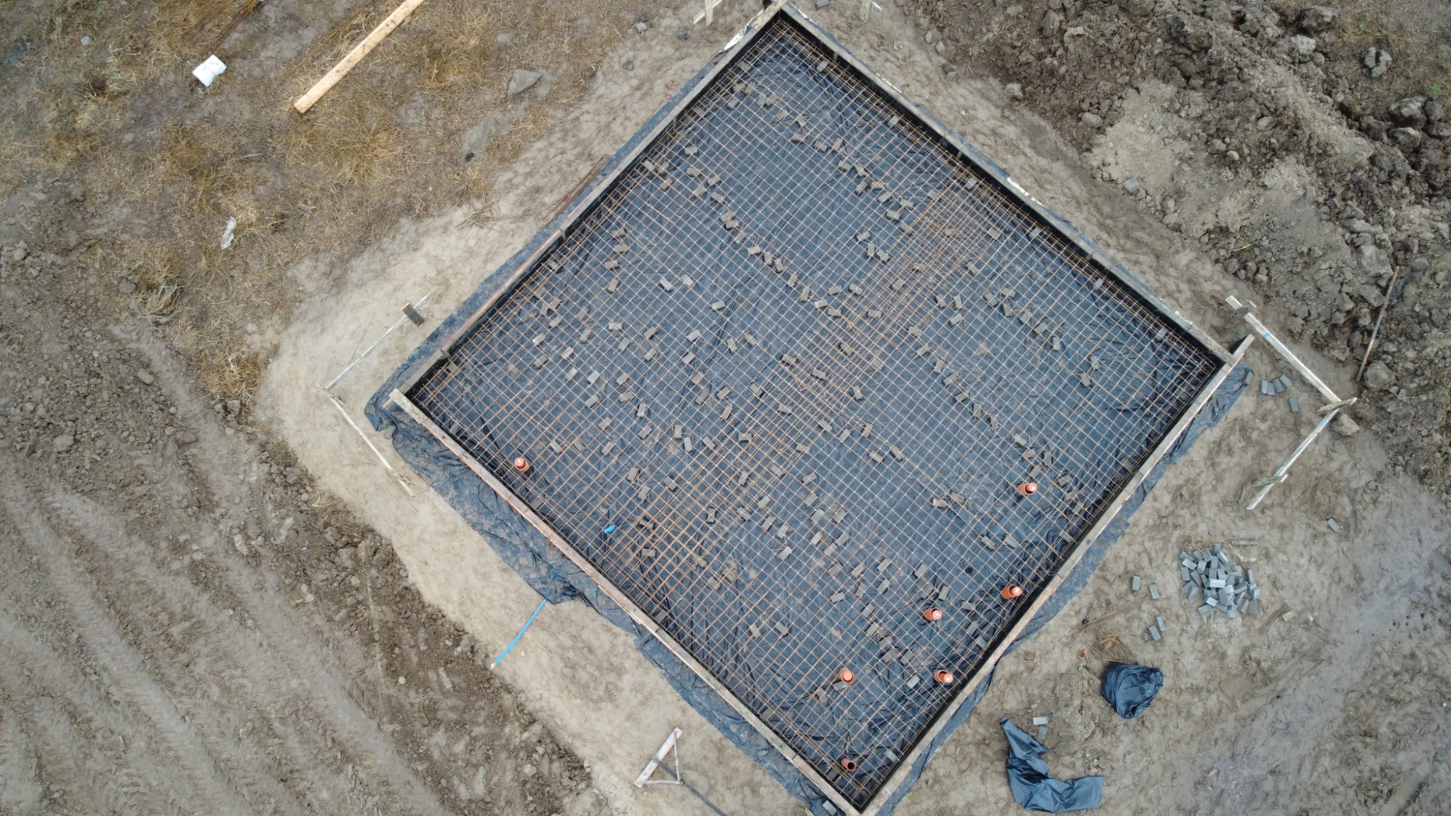 Widok z góry na przygotowany zbrojony fundament pod budynek, z deskowaniem, folią, siatką zbrojeniową podpartą kostkami dystansowymi i kilkoma pomarańczowymi rurami