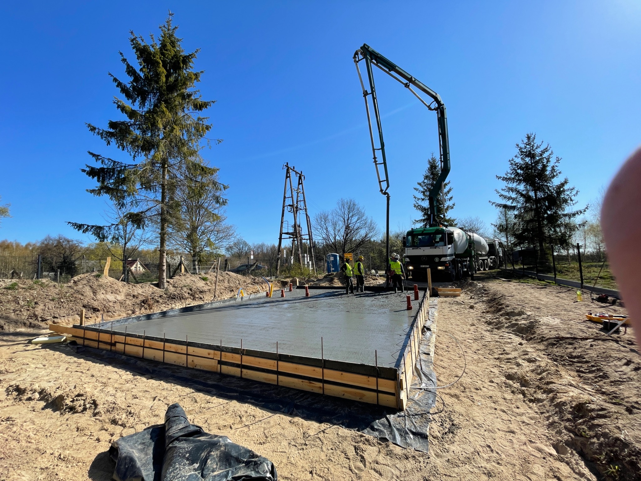 Wylewanie betonowej płyty fundamentowej pod budowę, widoczny betonowóz z pompą, szalunek z desek, pracownicy w kamizelkach odblaskowych i hełmach, teren budowy z piaskiem i drzewami, błękitne niebo.
