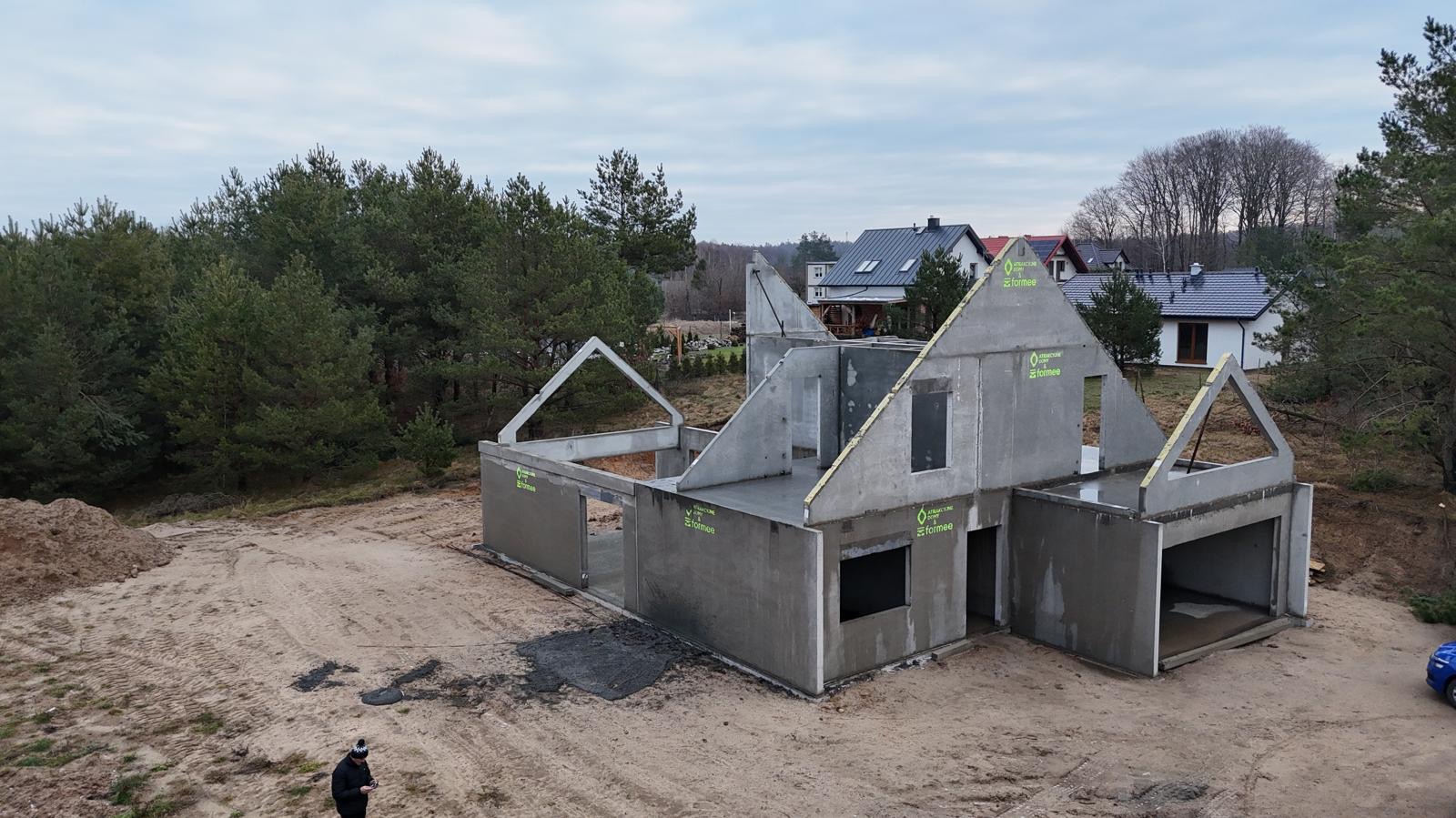 Surowy stan domu z prefabrykatów betonowych na działce z widocznymi drzewami i innymi domami w tle. Widoczny fragment niebieskiego samochodu.