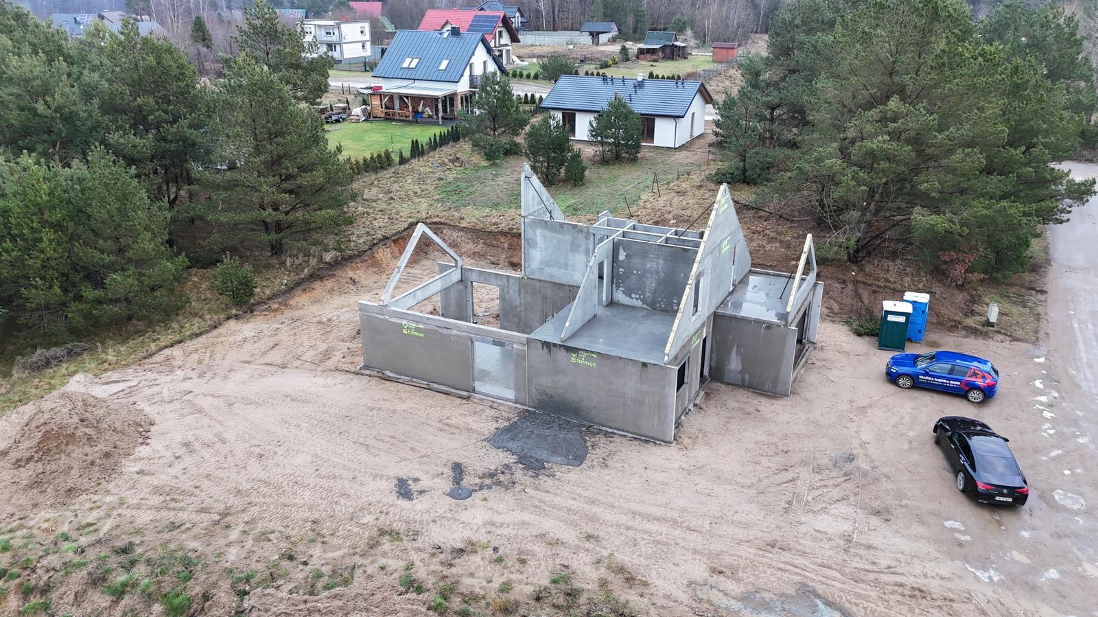 Surowy stan domu z prefabrykatów betonowych na działce, widok z góry. Na posesji zaparkowane dwa samochody osobowe i toaleta przenośna.