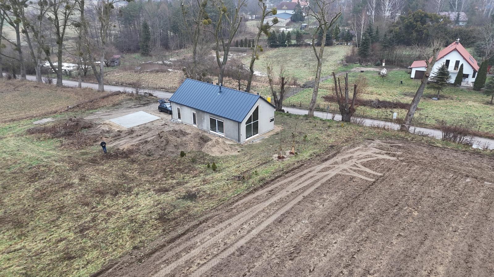 Nowoczesny, parterowy dom z szarymi ścianami i ciemnym dachem na działce z przygotowanym podjazdem, widziany z góry. W tle drzewa i zabudowania.