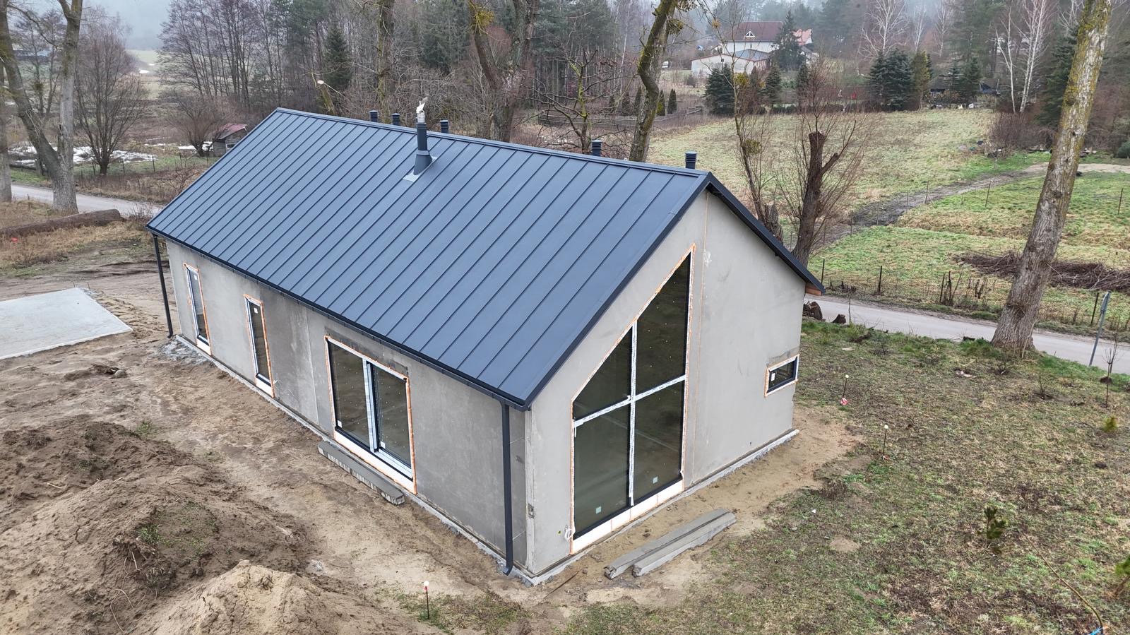 Surowy dom jednorodzinny z ciemnym dachem na działce, widok z góry. Duże okno z białymi szprosami. Budynek w stanie deweloperskim, otoczony ziemią i drzewami.