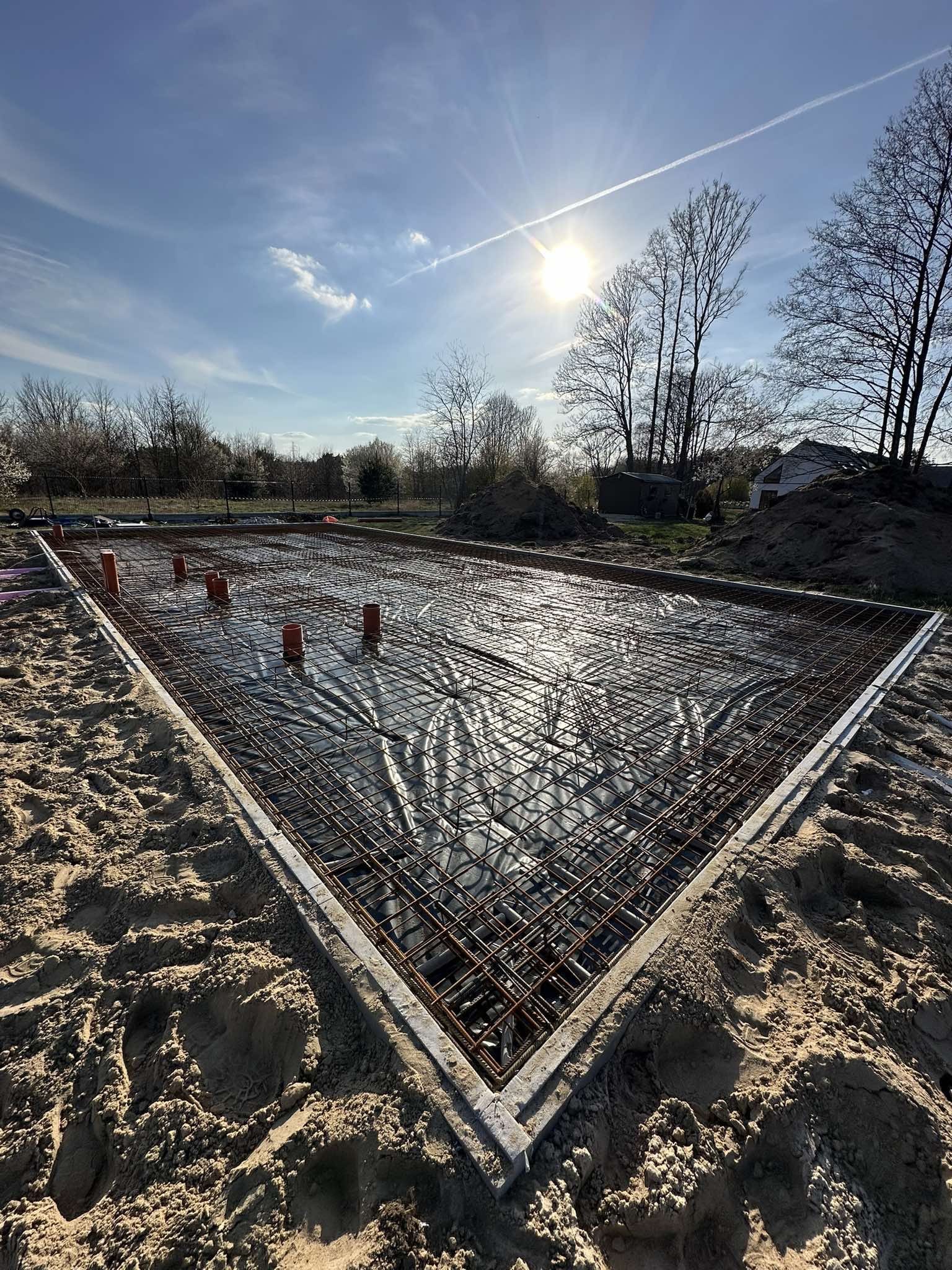 Zbrojenie fundamentów pod budowę domu, widok z góry na siatkę zbrojeniową na folii, z wystającymi rurami, otoczone piaskiem i betonową ramą. Słoneczny dzień.