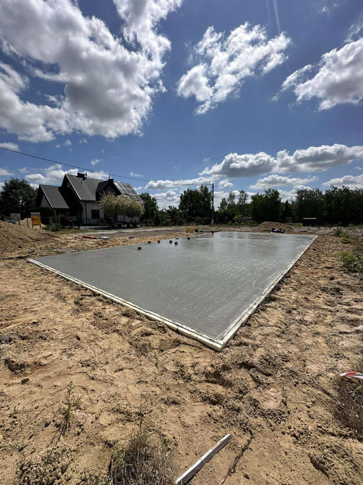 Świeżo wylana, gładka płyta fundamentowa pod budowę domu jednorodzinnego, otoczona piaskiem i z widocznym domem w tle, w słoneczny dzień.