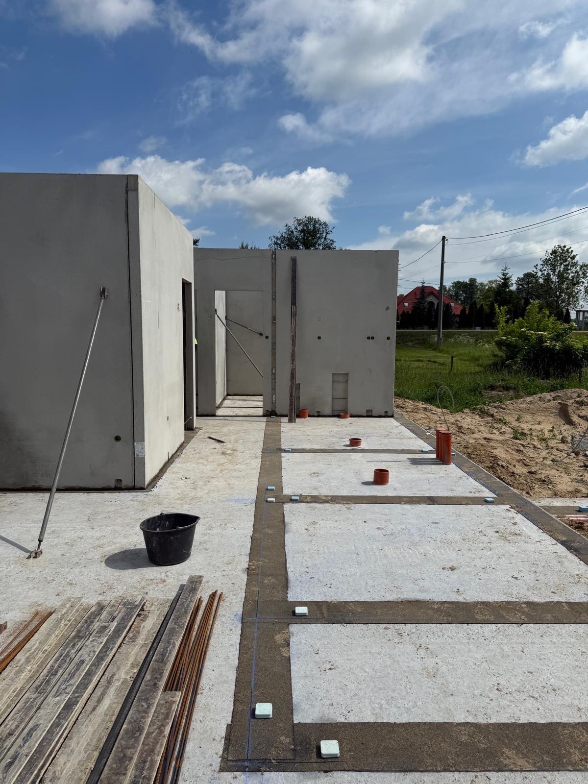 Wnętrze domu modułowego w trakcie budowy: widoczne betonowe ściany, fundamenty z izolacją, rury kanalizacyjne i narzędzia budowlane. Słoneczny dzień, błękitne niebo.