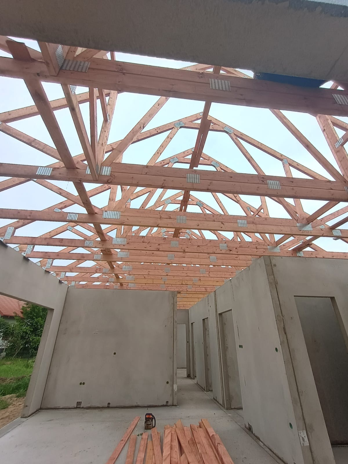 Wnętrze domu w trakcie budowy: surowe ściany z betonu i drewniana konstrukcja dachu z metalowymi łącznikami. Na podłodze leżą deski i piła spalinowa.