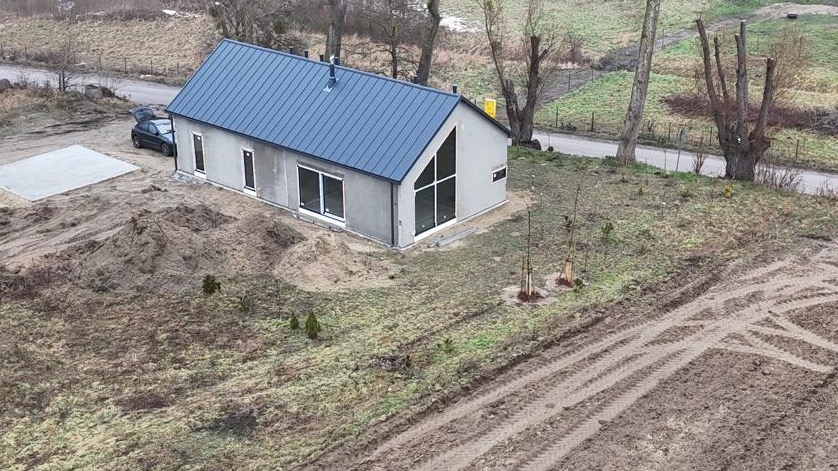 Nowoczesny, parterowy dom z szarą blachodachówką, dużym oknem trójkątnym i przygotowanym betonowym podłożem, otoczony ziemią z widocznymi śladami opon.