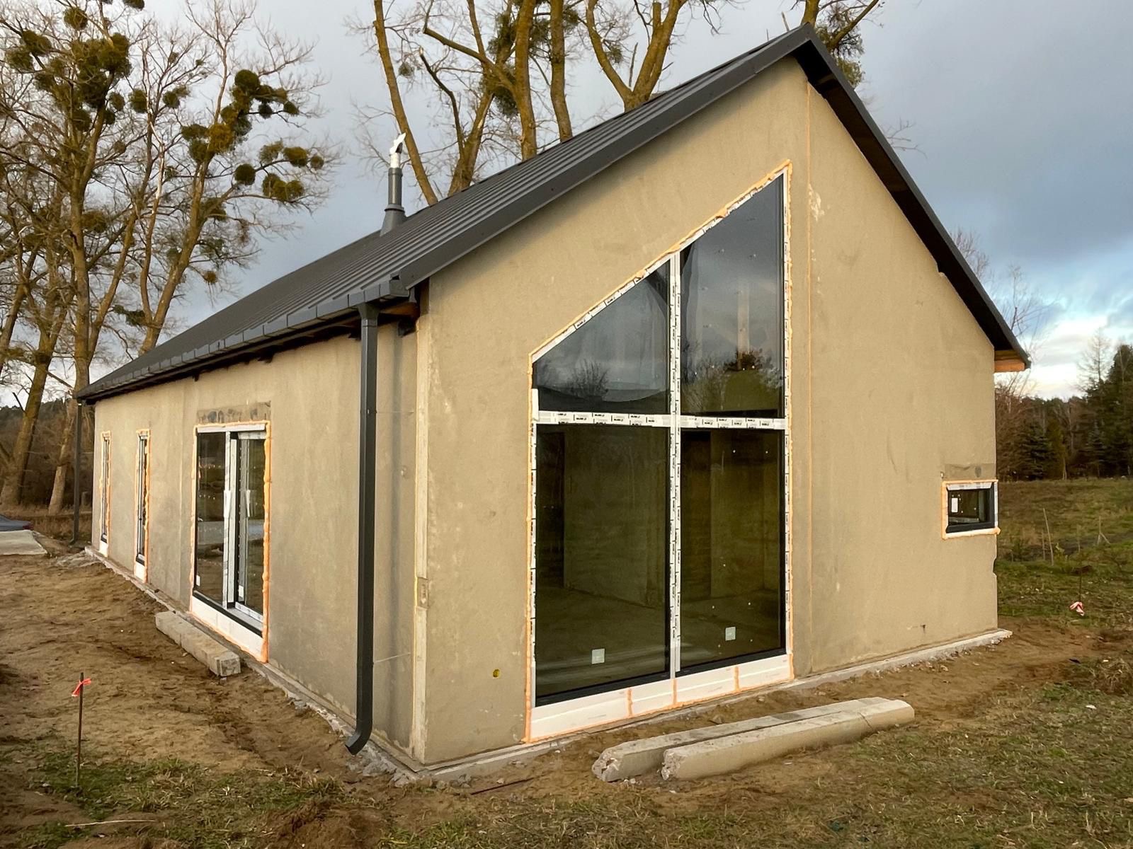 Surowy dom z dużym, trójkątnym oknem i szarą blachodachówką na tle drzew. Elewacja w trakcie prac wykończeniowych, widoczne zabezpieczenia okien.