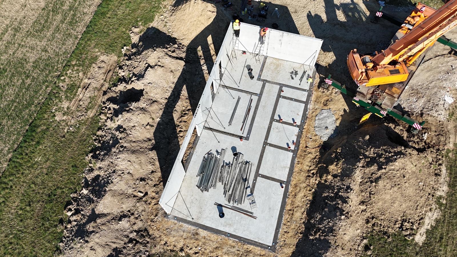 Fundament domu z prefabrykatów betonowych, widok z góry. Na fundamencie widoczne elementy konstrukcyjne i pracownicy w kaskach. Żuraw budowlany w tle.