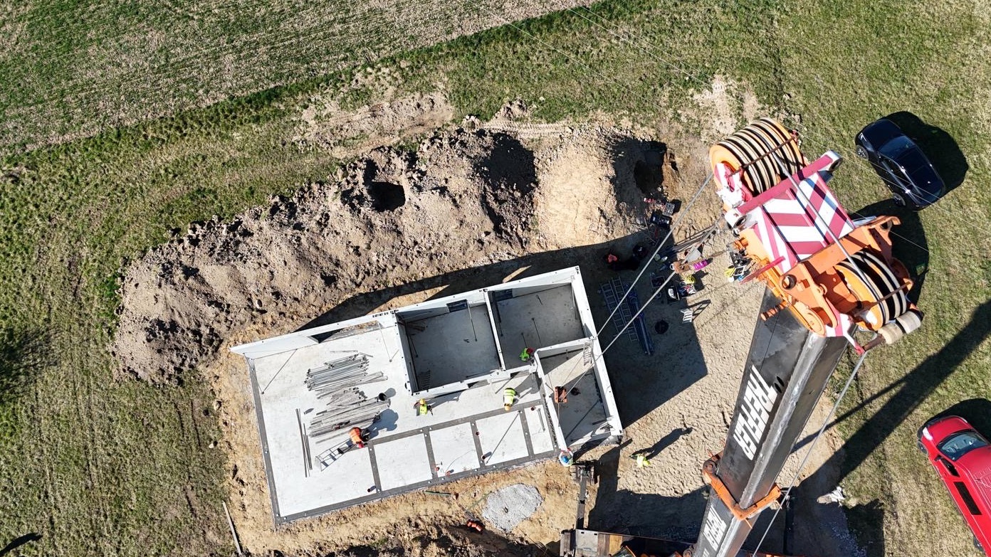 Widok z góry na fundamenty domu w trakcie budowy, z dźwigiem i pracownikami w kaskach, otoczone ziemią i trawą. W tle zaparkowane samochody.