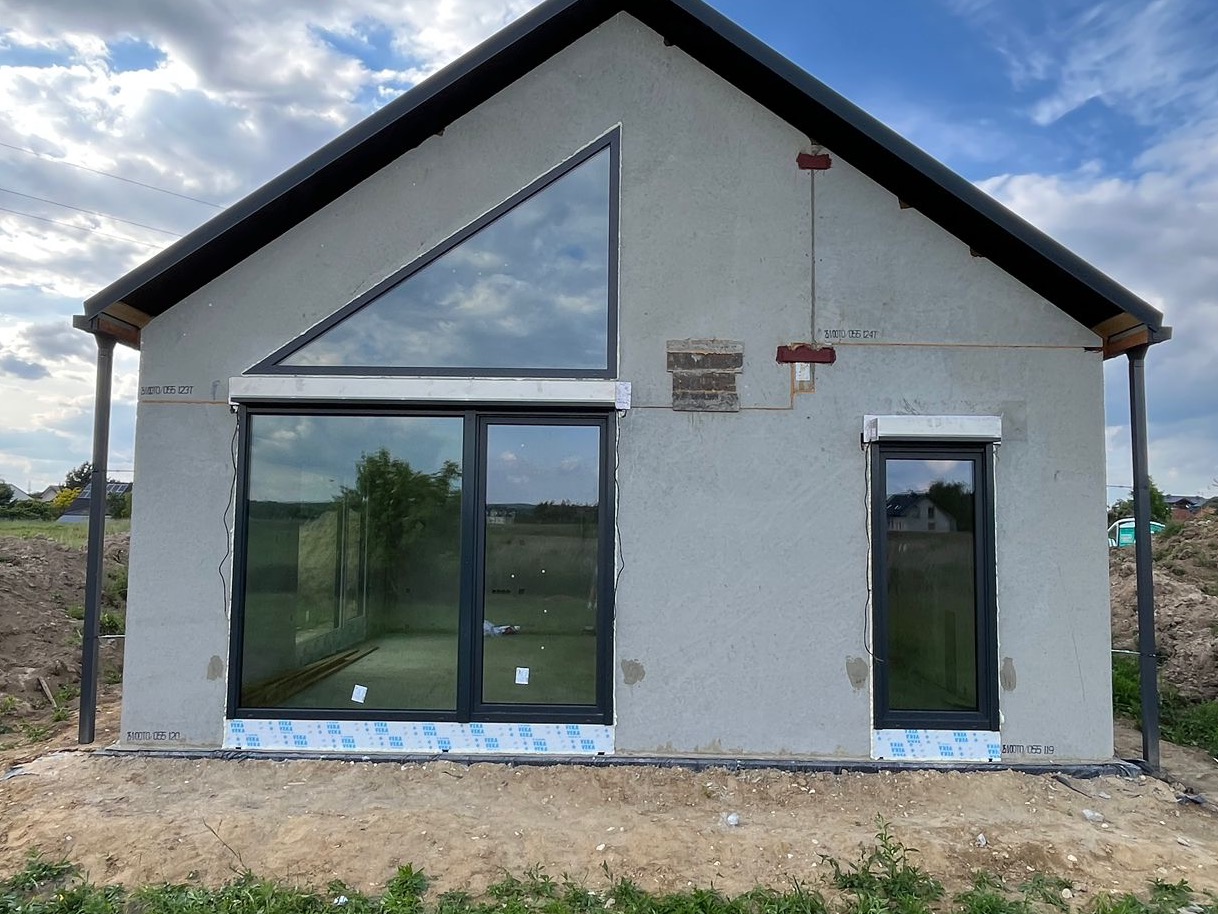 Surowy stan domu z dużym trójkątnym oknem i przesuwnymi drzwiami tarasowymi, widoczne instalacje na elewacji, teren wokół domu w trakcie prac ziemnych.