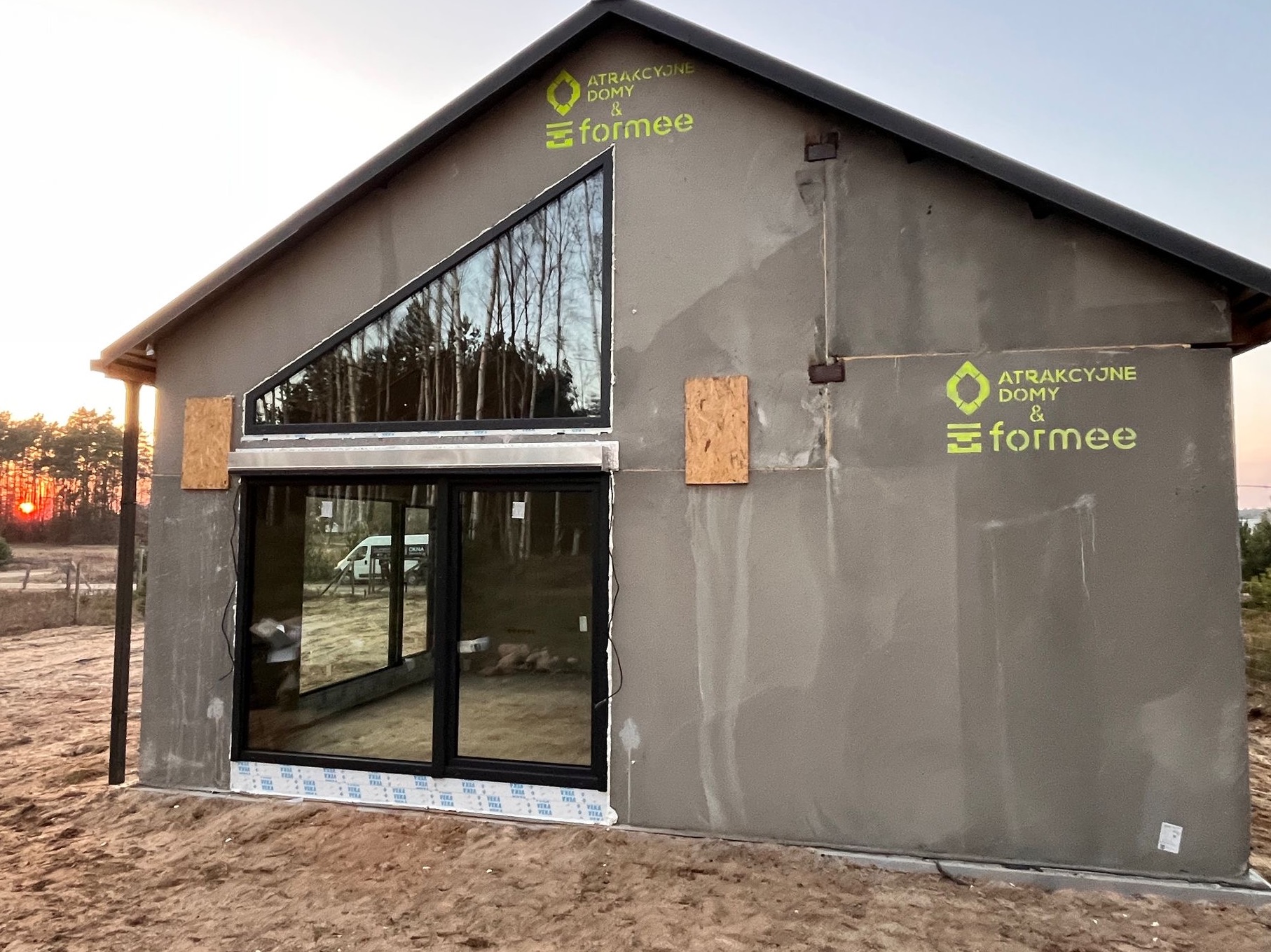 Surowy, otynkowany dom jednorodzinny z dużymi oknami, w tym trójkątnym na szczycie. Widoczne logo firmy 'Atrakcyjne Domy & Formee' na elewacji. Zachód słońca w tle.