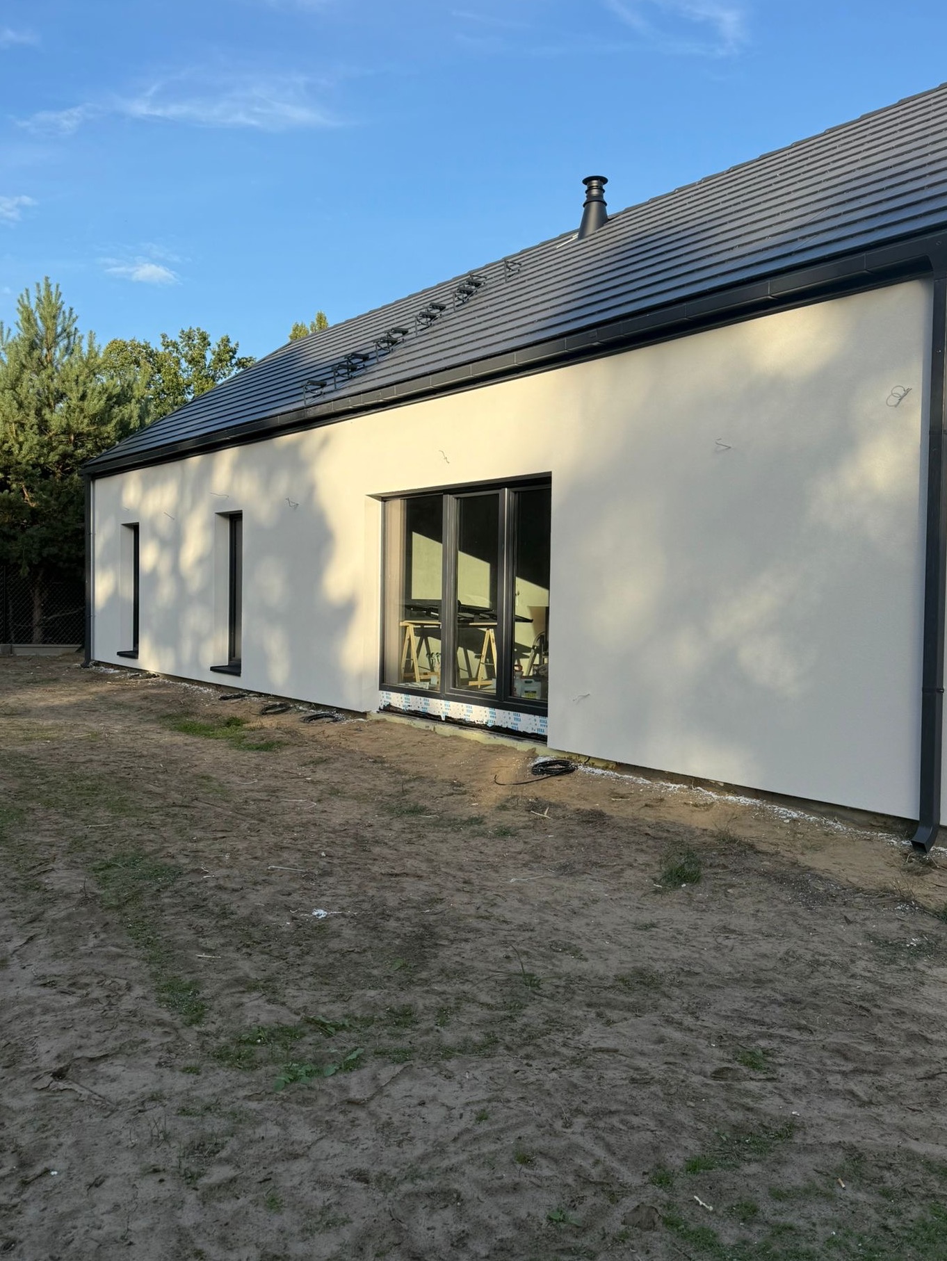 Nowoczesny dom jednorodzinny z jasną elewacją i ciemną dachówką, widok z boku na tle błękitnego nieba i zieleni drzew. Okna z czarnymi ramami.