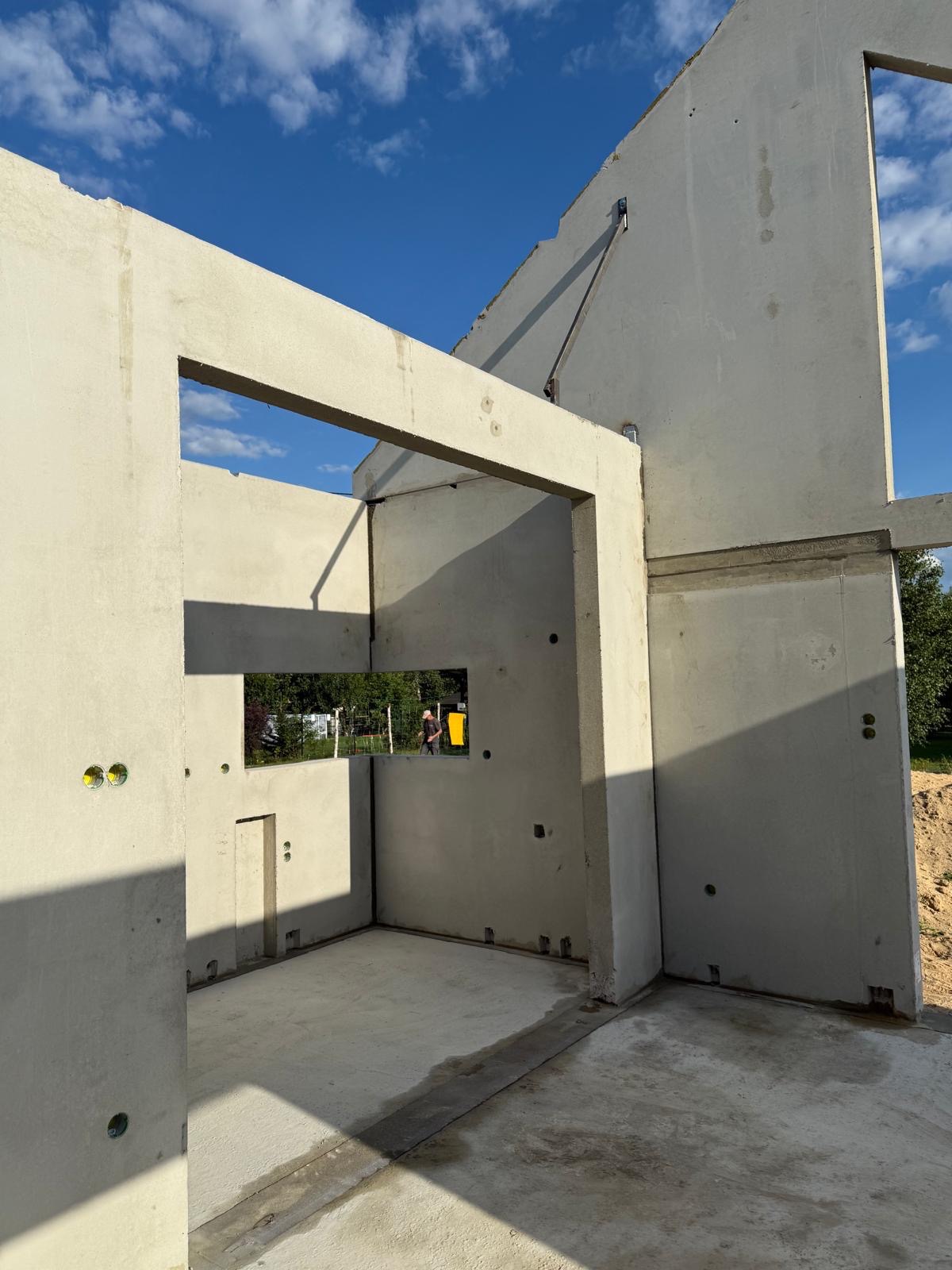 Surowy stan domu z prefabrykatów betonowych w słoneczny dzień. Widoczne otwory okienne i drzwiowe, ślady po szalunkach i elementy złączne. W tle zieleń.