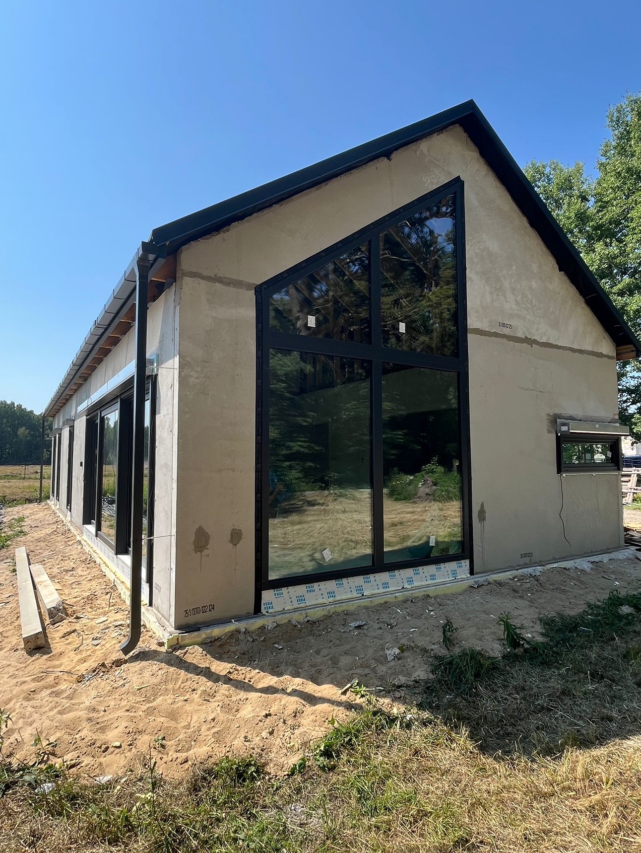 Surowy dom jednorodzinny z dużymi, czarnymi oknami i szarą elewacją w trakcie budowy, otoczony piaskiem i zielenią, w słoneczny dzień.