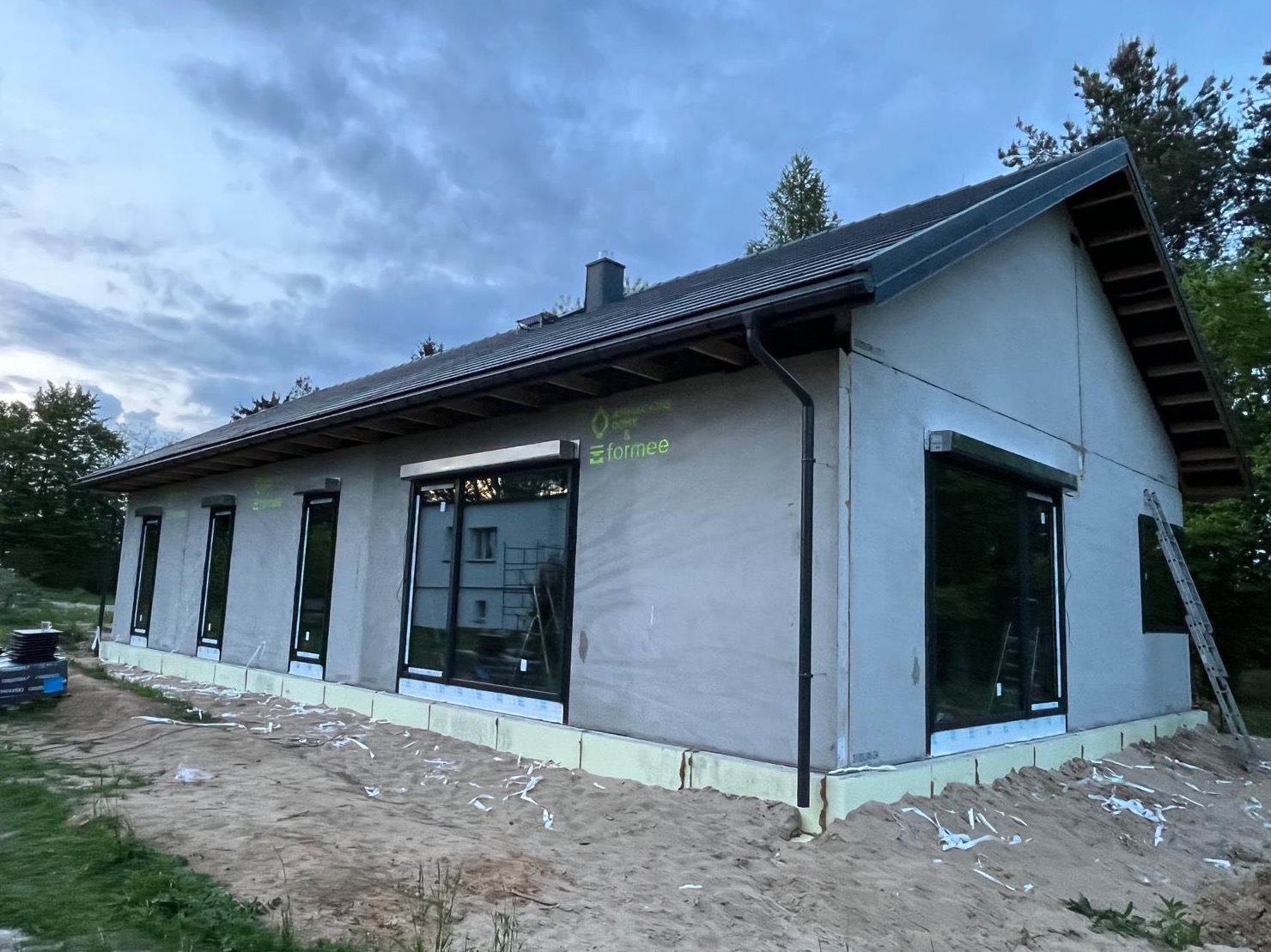 Niski dom parterowy z szarą elewacją w trakcie budowy, z widocznymi oknami z roletami zewnętrznymi i ociepleniem fundamentów. Na elewacji widoczne logo producenta.