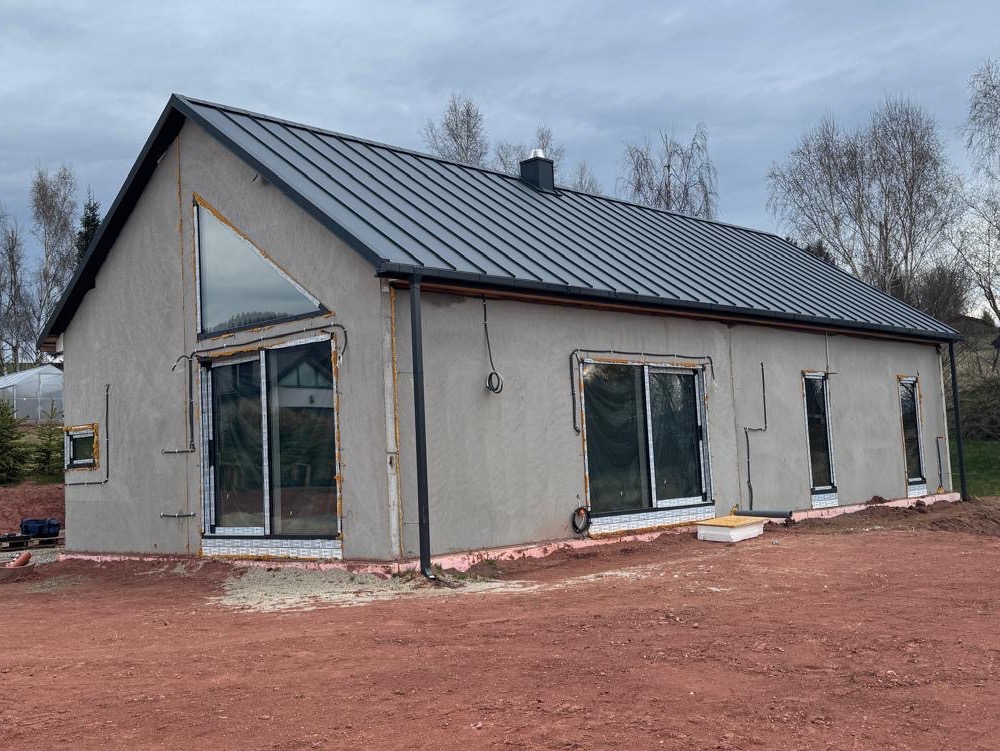 Surowy, parterowy dom z szarą blachodachówką i dużymi oknami, w trakcie wykończenia elewacji. Widoczne ocieplenie wokół okien i czerwona ziemia wokół budynku.