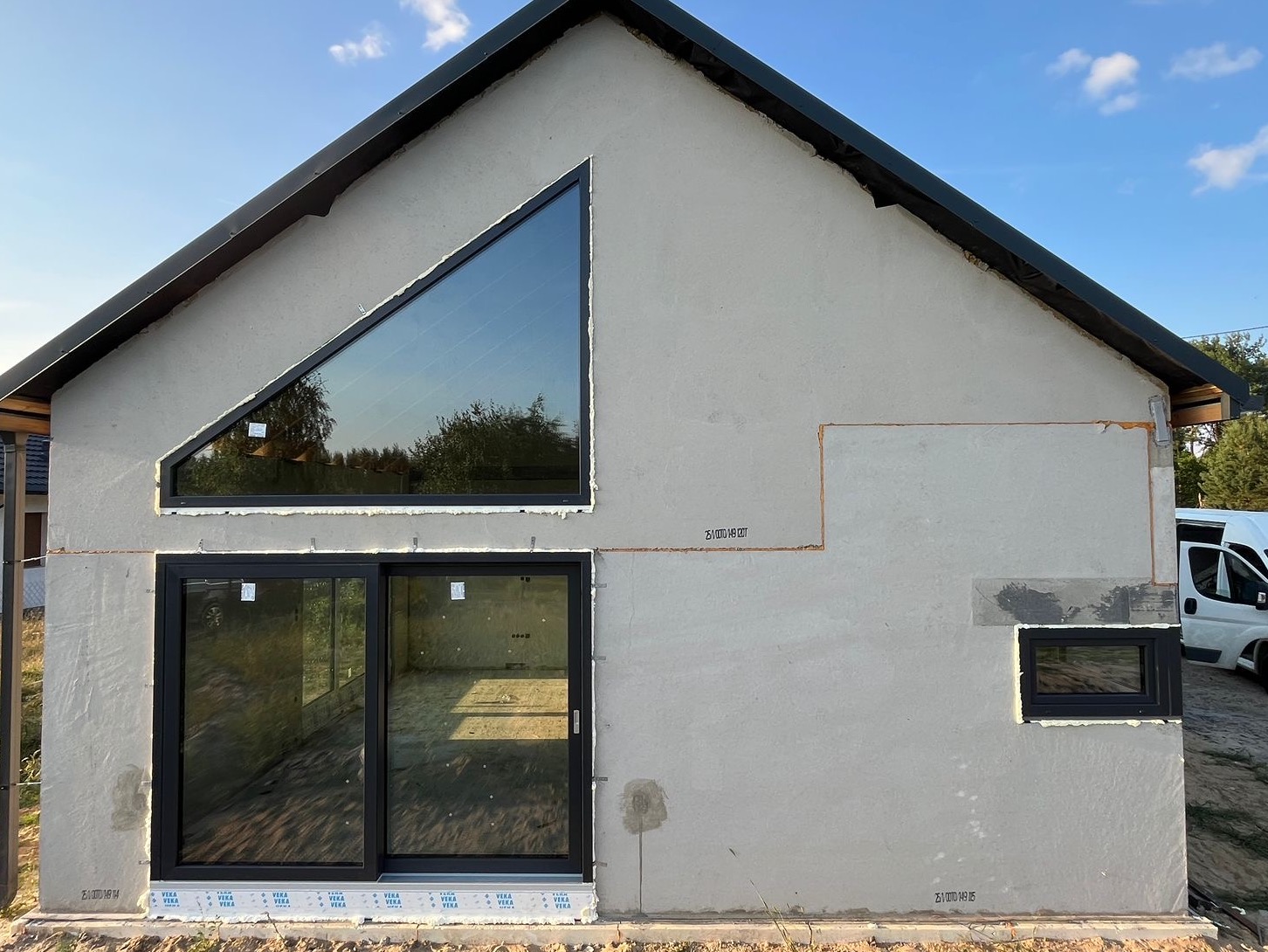 Niedokończony dom z oknem trójkątnym i przesuwnymi drzwiami balkonowymi. Widoczne ślady po łatach na elewacji i brak wykończenia.