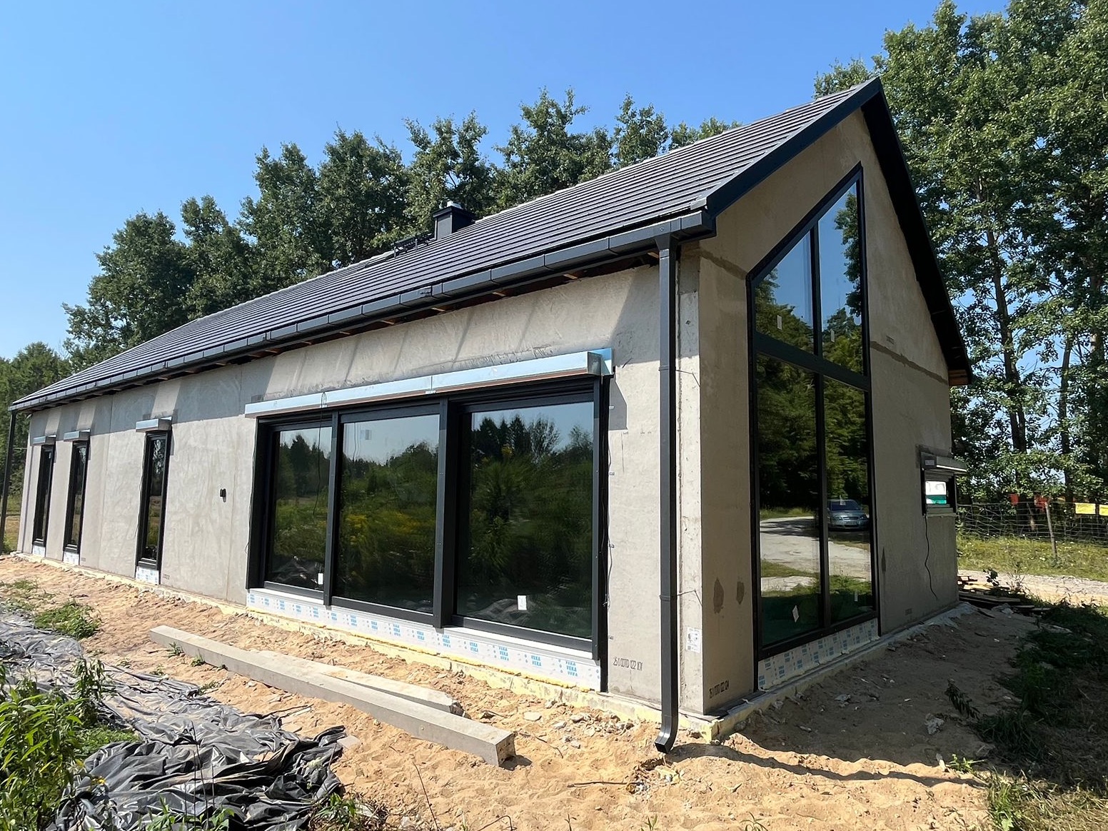 Surowy dom jednorodzinny z dużymi oknami i szarą dachówką, w trakcie wykończenia elewacji. Widoczne przygotowania do dalszych prac budowlanych.