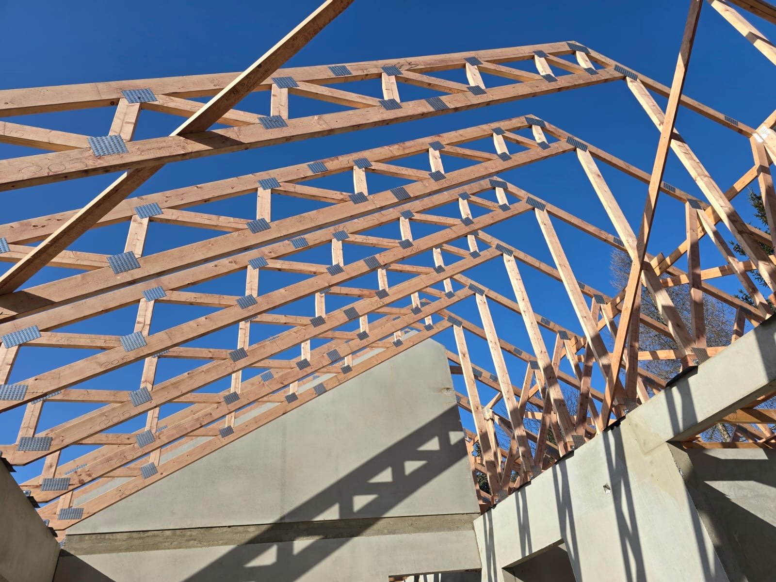 Konstrukcja drewniana dachu z metalowymi łącznikami na tle błękitnego nieba, widok z dołu. Częściowo widoczne ściany budynku w surowym stanie.