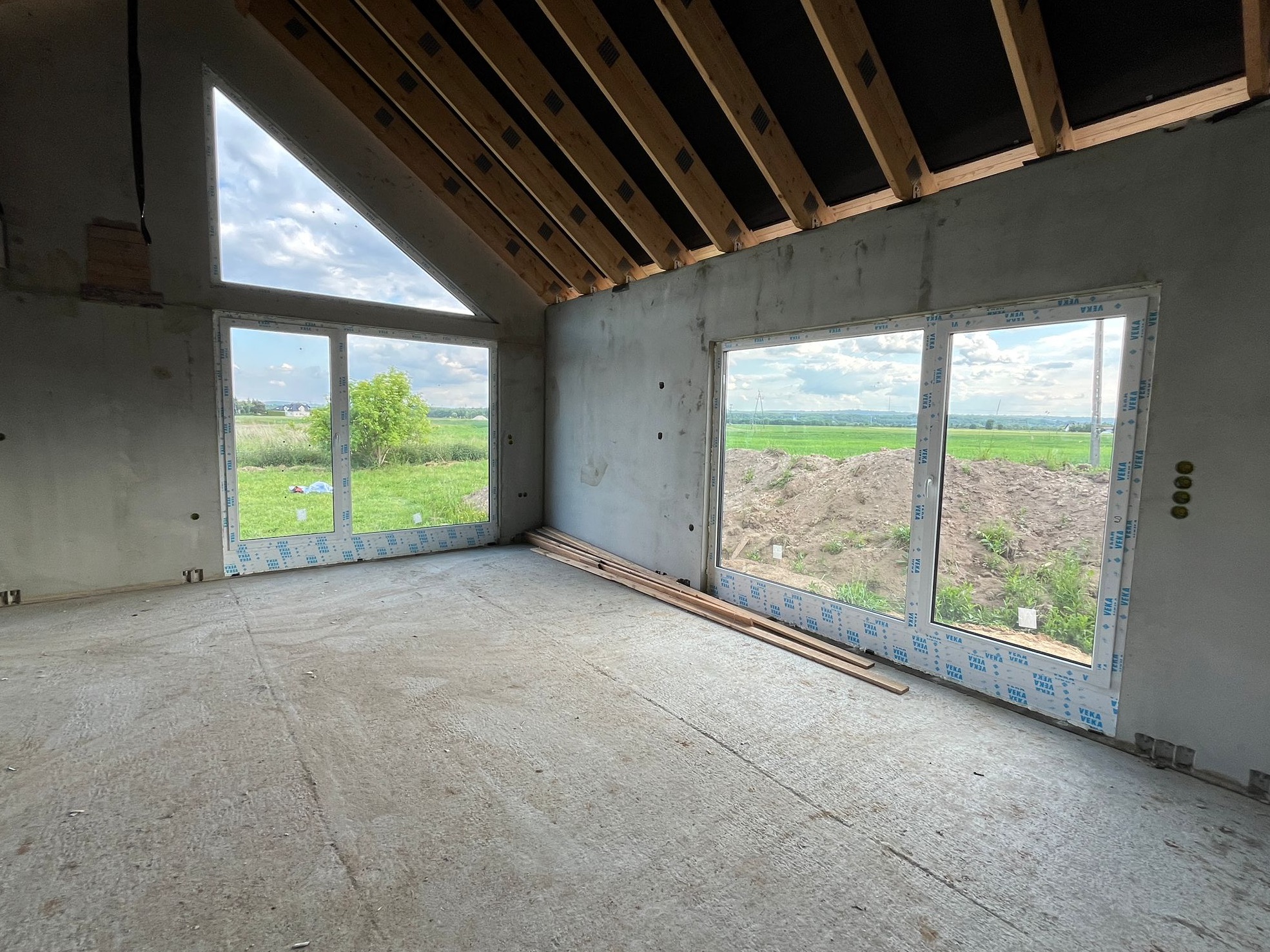 Surowy stan wnętrza domu w budowie: widoczne okna z folią ochronną, drewniane belki stropowe i surowe ściany. Na podłodze leżą deski.