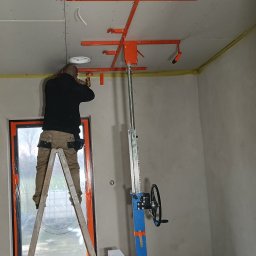 PHU COMPLEX GRZEGORZ ZIMNIAK - Montaż płyty gipsowej na suficie za pomocą podnośnika. Pracownik na drabinie przykręca płytę. Widoczne narzędzia i fragment okna. Żółta taśma na łączeniu ściany z sufitem.