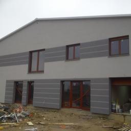 PHU COMPLEX GRZEGORZ ZIMNIAK - Dwukondygnacyjny budynek z szarą elewacją i poziomymi pasami, widoczne okna z drewnianymi ramami, teren wokół budynku z gruzem i materiałami budowlanymi.