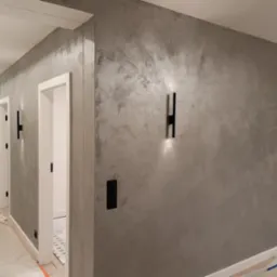 Wnętrze korytarza z dekoracyjną szarą ścianą o fakturze betonu, oświetloną przez nowoczesne, czarne kinkiety, widoczne drzwi z czarnymi klamkami i osprzęt elektryczny.